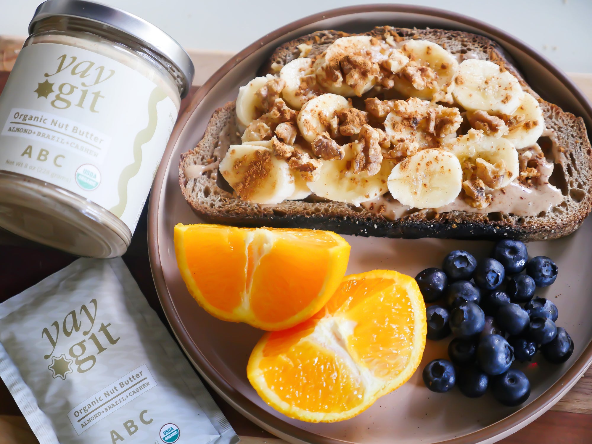 Toast with banana slices, blueberries, and orange slices on a plate with yaygit nut butter jar and packets.